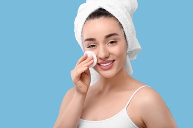 Photo of Beautiful woman in terry towel removing makeup with cotton pad on light blue background