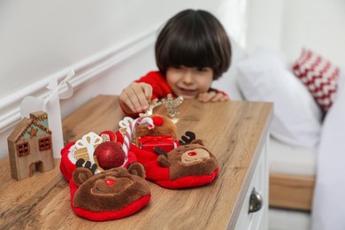 Photo of Cute little boy near cabinet at home, focus on slippers with treats. Saint Nicholas day tradition