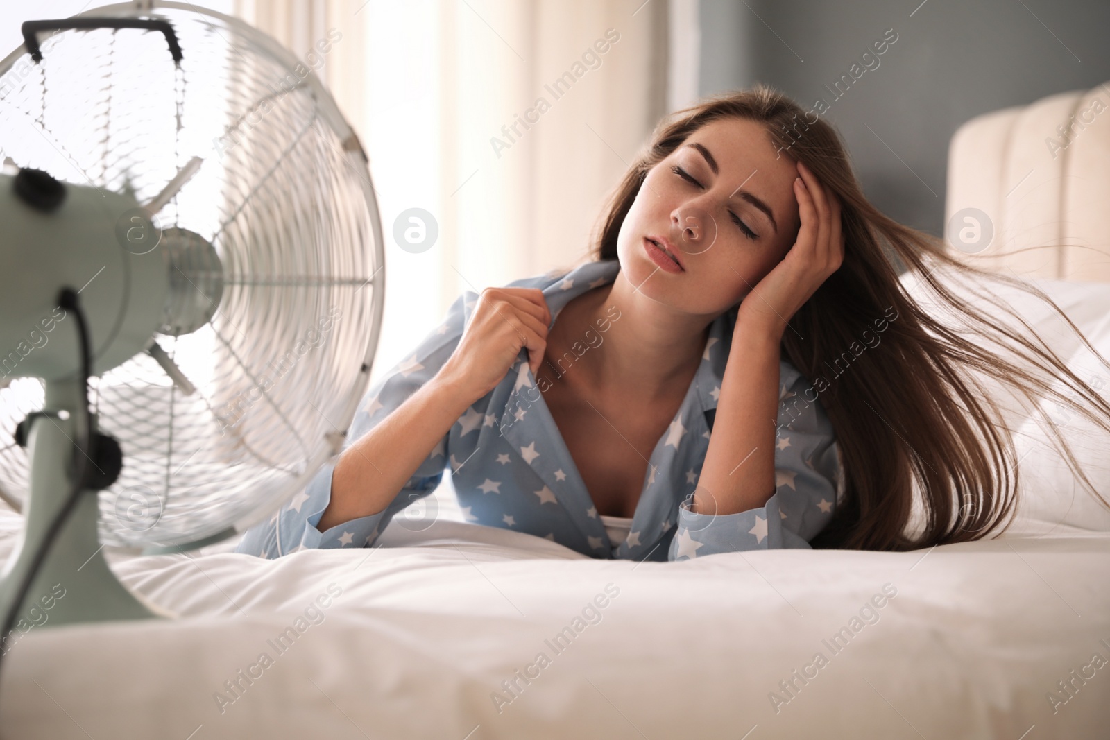 Photo of Woman enjoying air flow from fan on bed in room. Summer heat