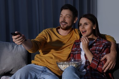 Photo of Happy couple watching TV at home in evening. Man changing channels with remote control