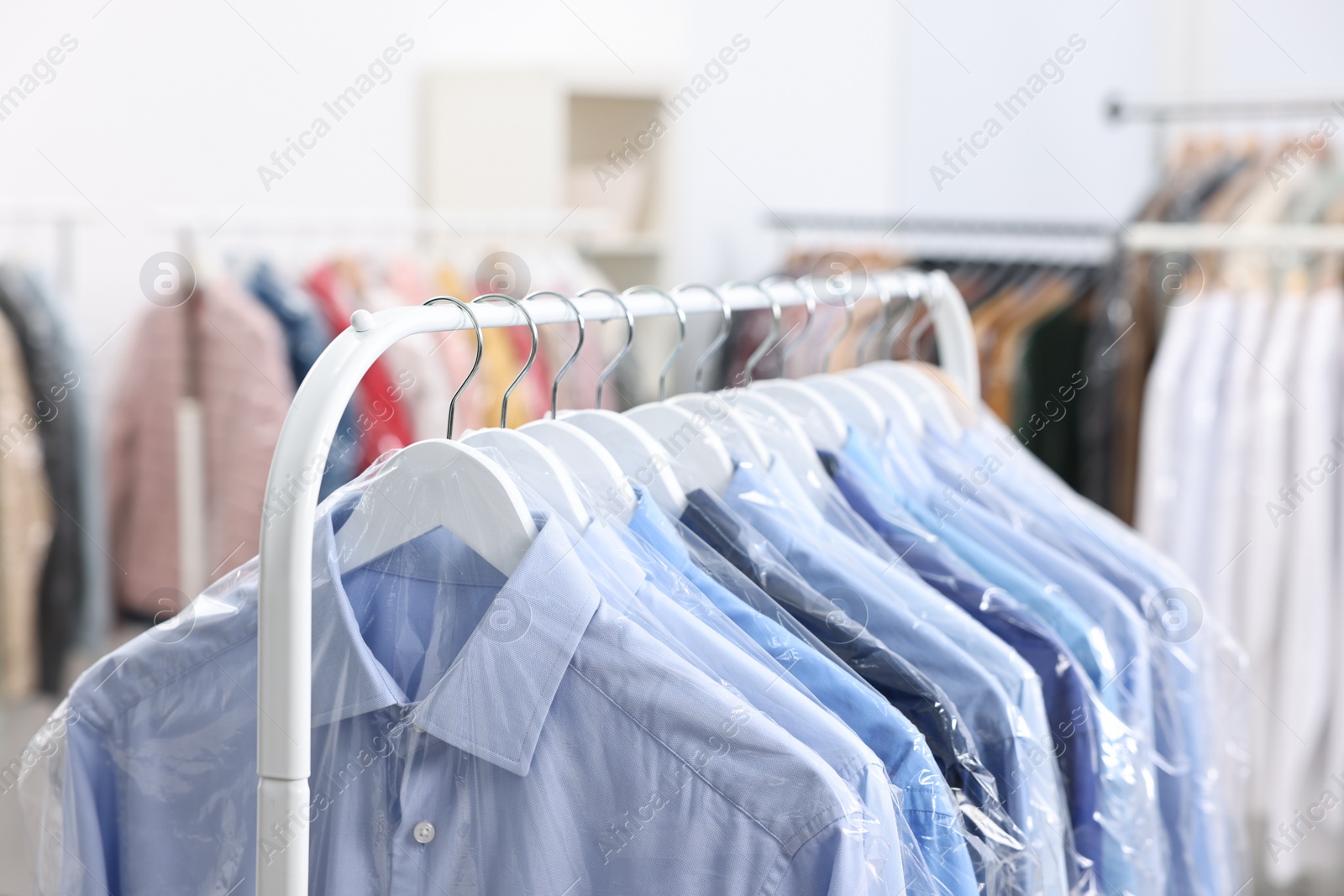 Photo of Dry-cleaning service. Many different clothes in plastic bags hanging on rack indoors, closeup