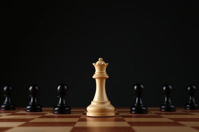 Photo of White queen among black pawns on wooden chess board against dark background