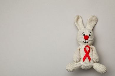 Photo of Cute knitted toy bunny with red ribbon on beige background, top view and space for text. AIDS disease awareness