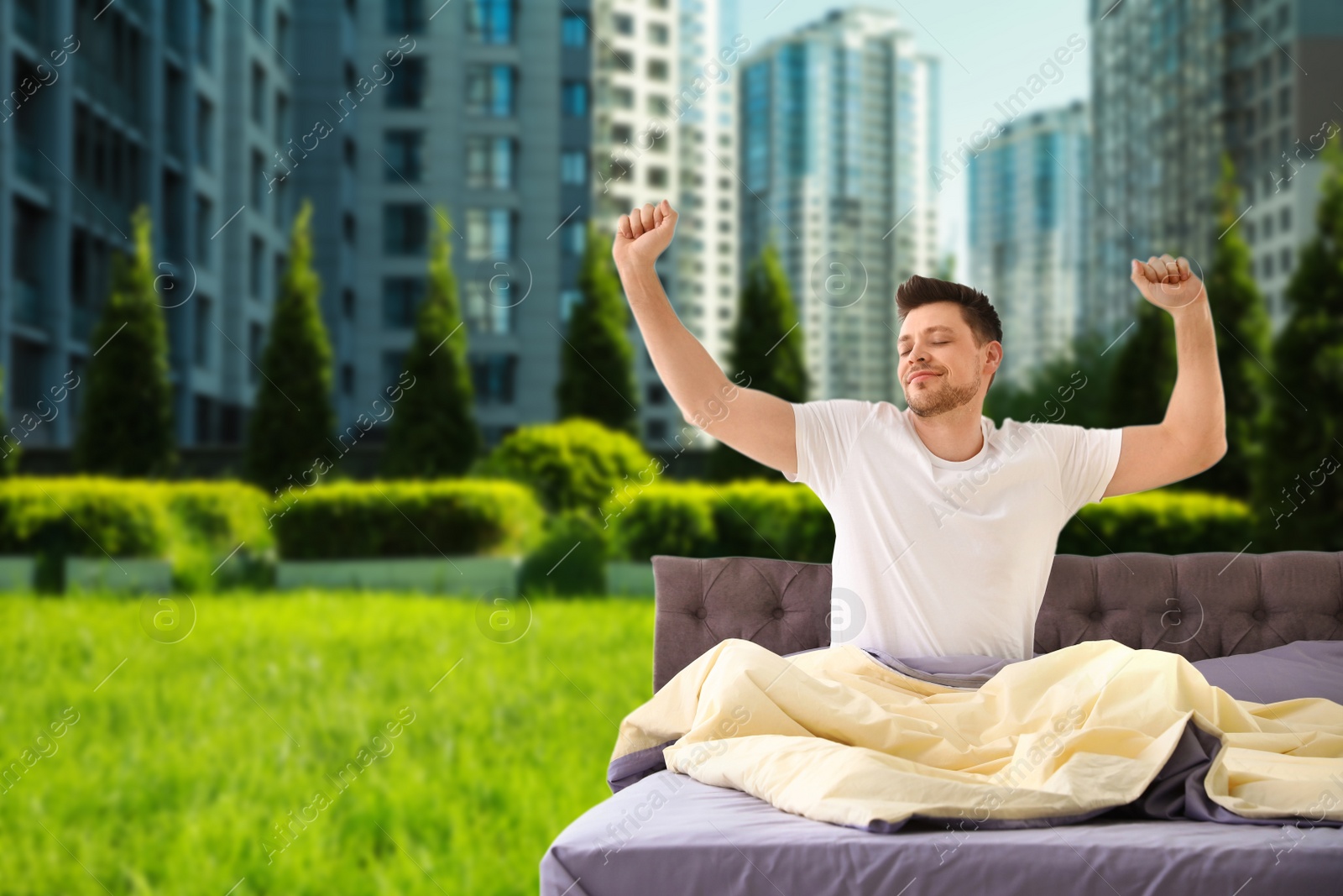 Image of Happy man stretching in bed and beautiful view of green grass and cityscape on background. Good sleep despite of urban noise