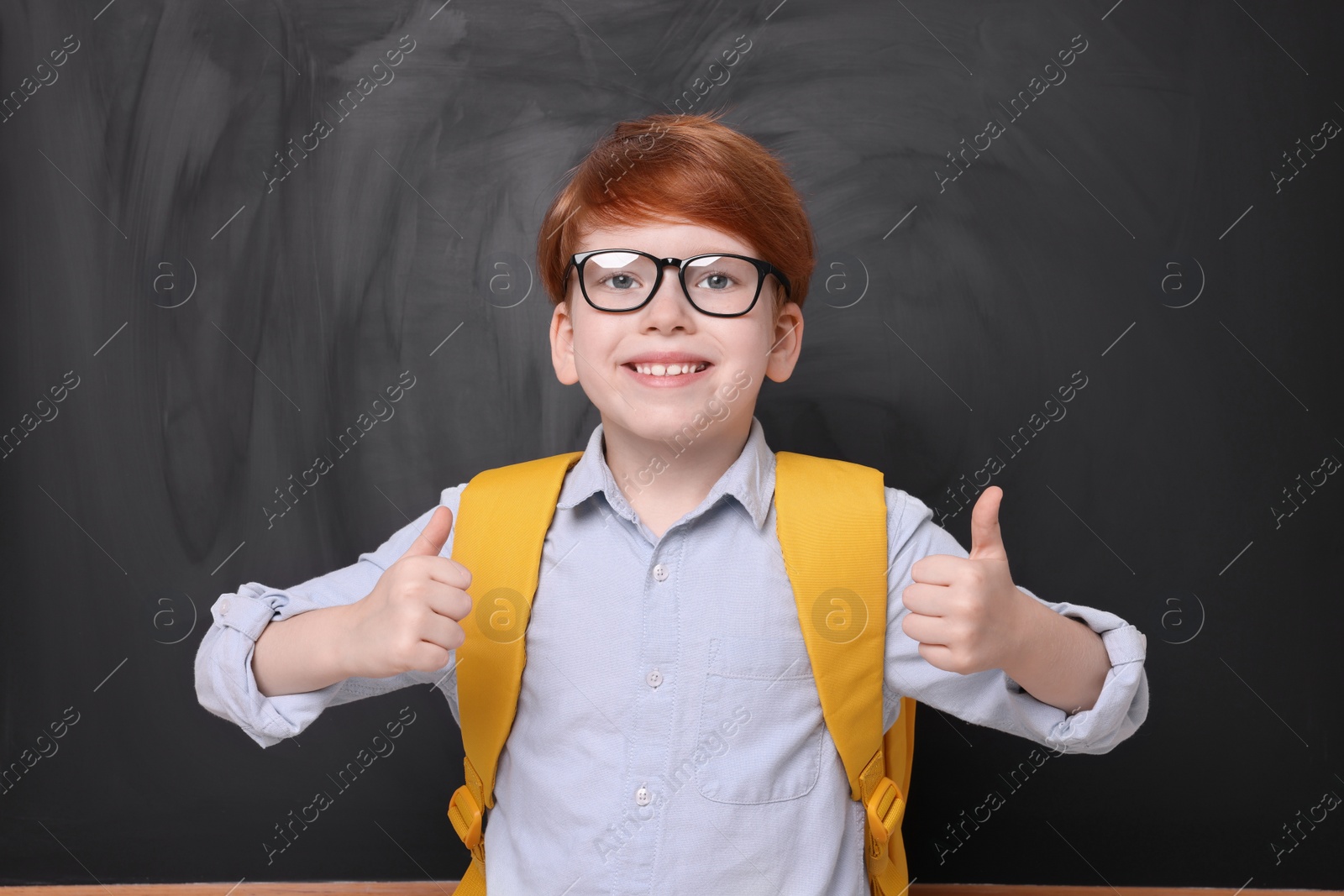 Photo of Smiling schoolboy in glasses showing thumbs up near blackboard