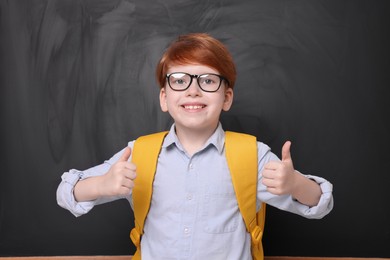 Photo of Smiling schoolboy in glasses showing thumbs up near blackboard
