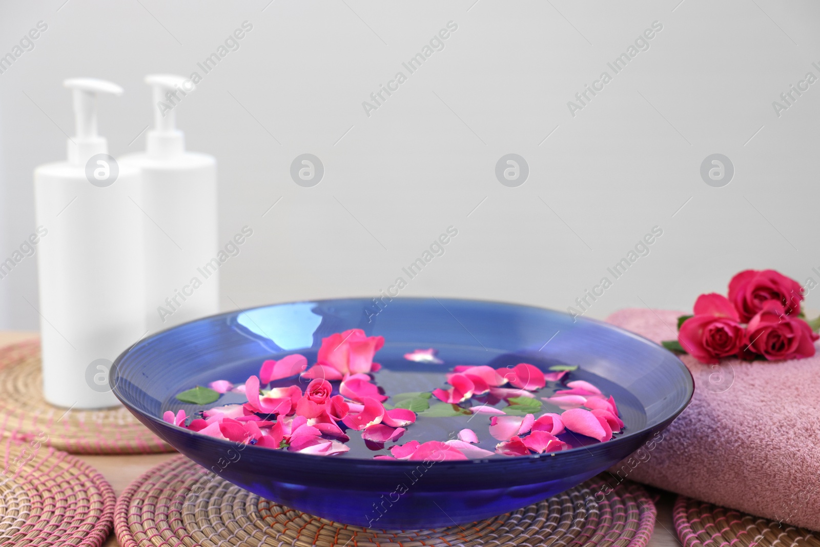 Photo of Pink roses, petals in bowl with water, cosmetic products and towel on table