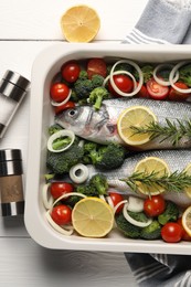 Raw fish with vegetables in baking dish and lemon on white wooden table, flat lay