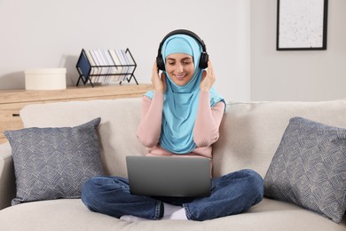 Muslim woman in headphones using laptop at couch in room