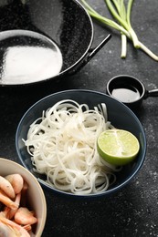 Photo of Noodles, lime and black wok on grey textured table, closeup