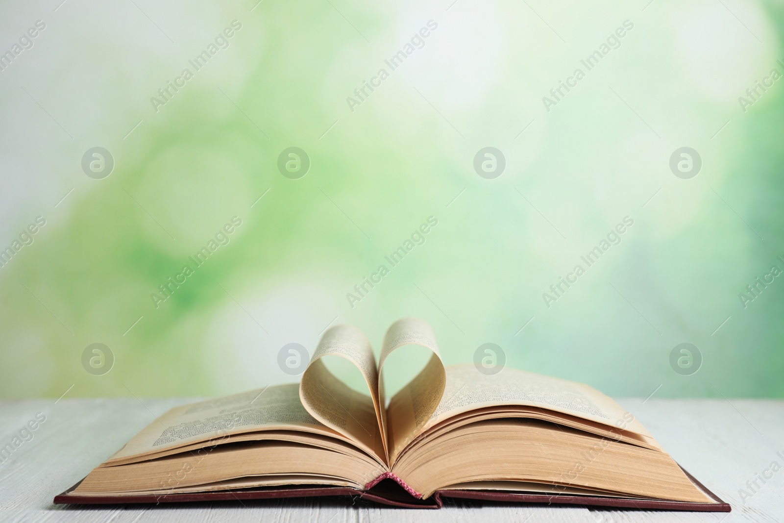 Photo of Open book on white wooden table against blurred green background. Space for text