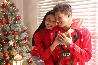 Photo of Happy couple in warm sweaters near Christmas tree indoors