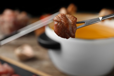 Fork with cooked meat and blurred fondue pot on background, closeup. Space for text