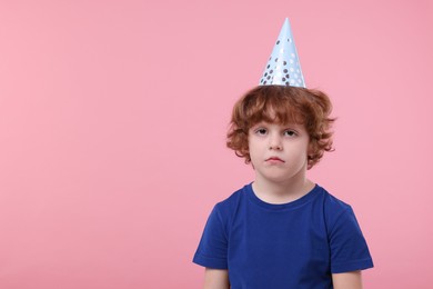 Upset little boy in party hat with blower on pink background. Space for text