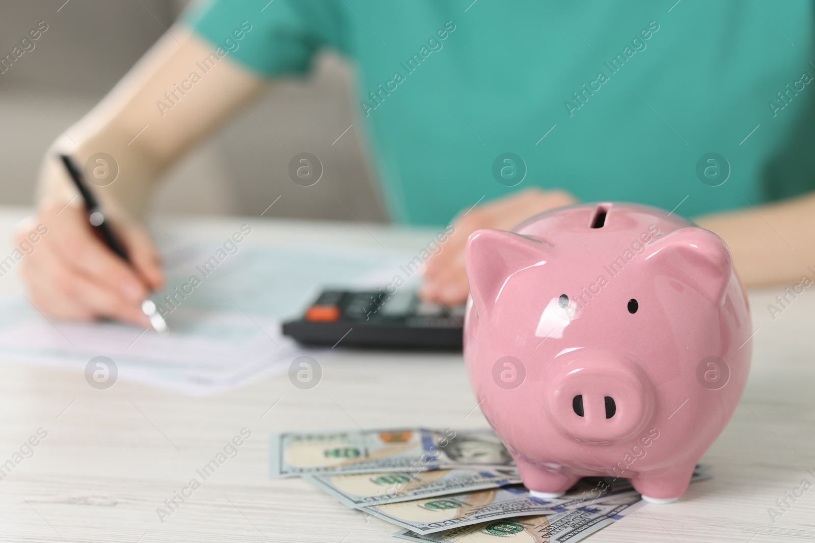 Photo of Financial savings. Woman using calculator at white wooden table indoors, focus on piggy bank and dollar banknotes