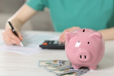 Photo of Financial savings. Woman using calculator at white wooden table indoors, focus on piggy bank and dollar banknotes