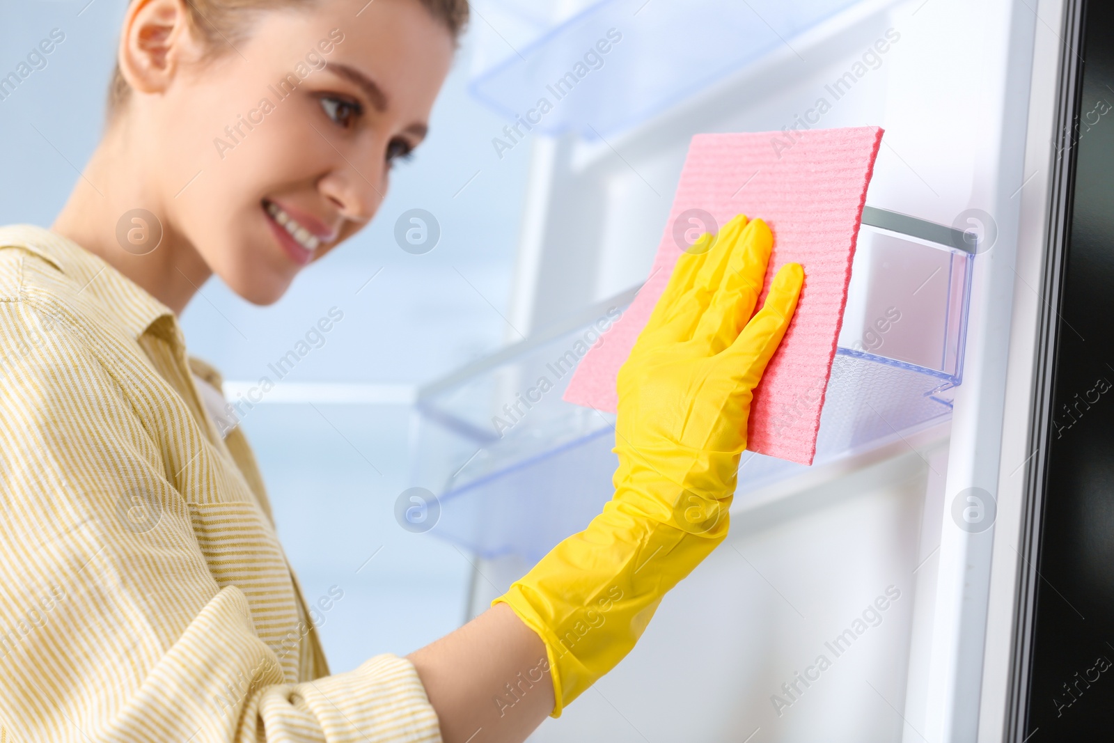 Photo of Woman in rubber gloves cleaning empty refrigerator at home, focus on hand with rag