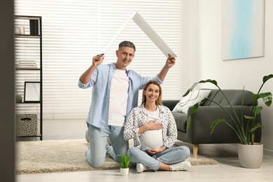 Photo of Young family housing concept. Man holding cardboard roof over his pregnant wife head at home