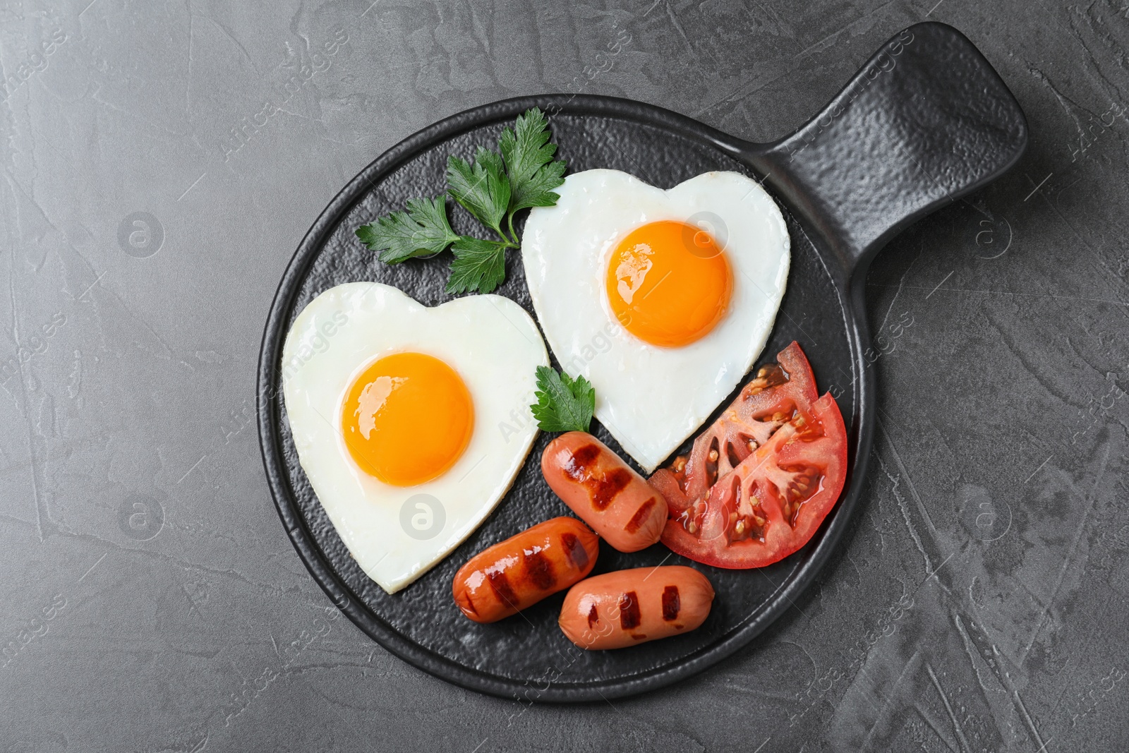 Photo of Slate plate of tasty breakfast with heart shaped fried eggs and sausages on dark grey table, top view