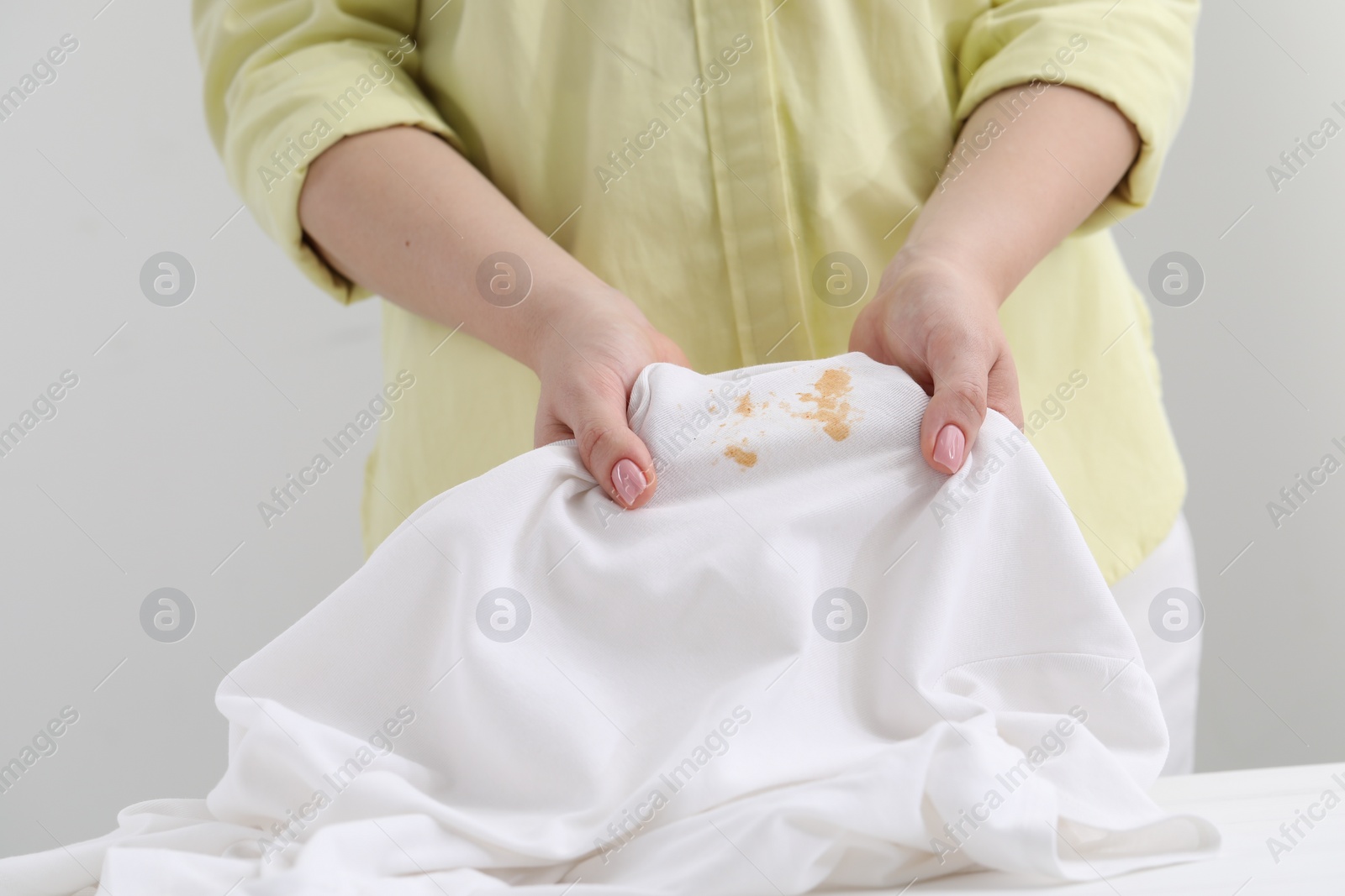 Photo of Woman holding shirt with stain at white table against light grey background, closeup