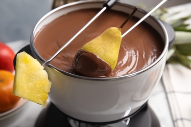 Photo of Dipping pear into pot with chocolate fondue on table, closeup