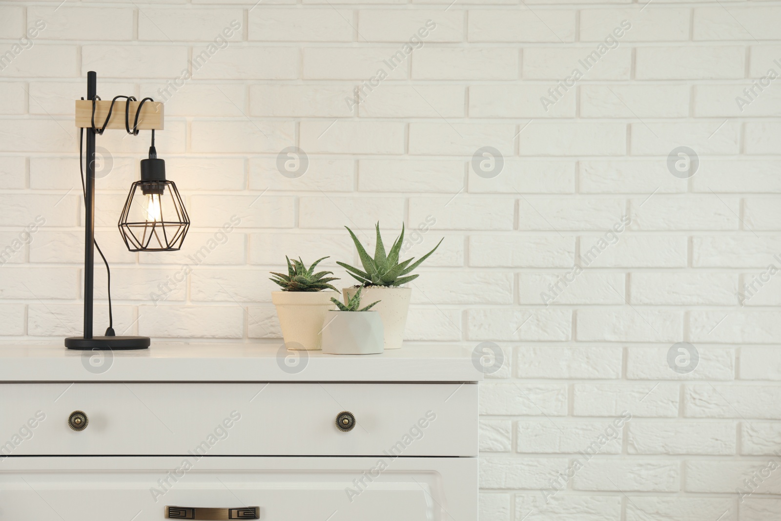 Photo of Modern chest of drawers with houseplants and lamp near white brick wall. Space for text