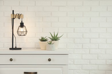 Modern chest of drawers with houseplants and lamp near white brick wall. Space for text