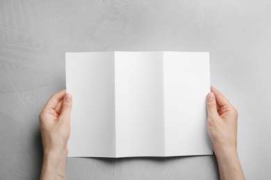 Young woman holding blank brochure at grey table, top view. Mock up for design