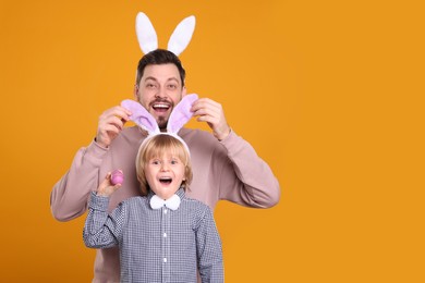 Photo of Father and son in bunny ears headbands having fun on orange background, space for text. Easter celebration