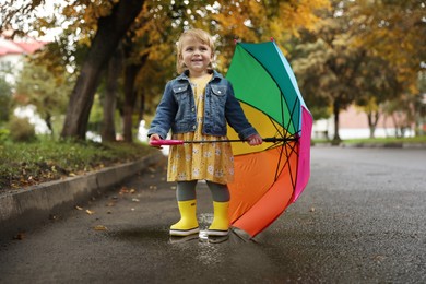 Photo of Cute little girl with colorful umbrella standing in puddle outdoors