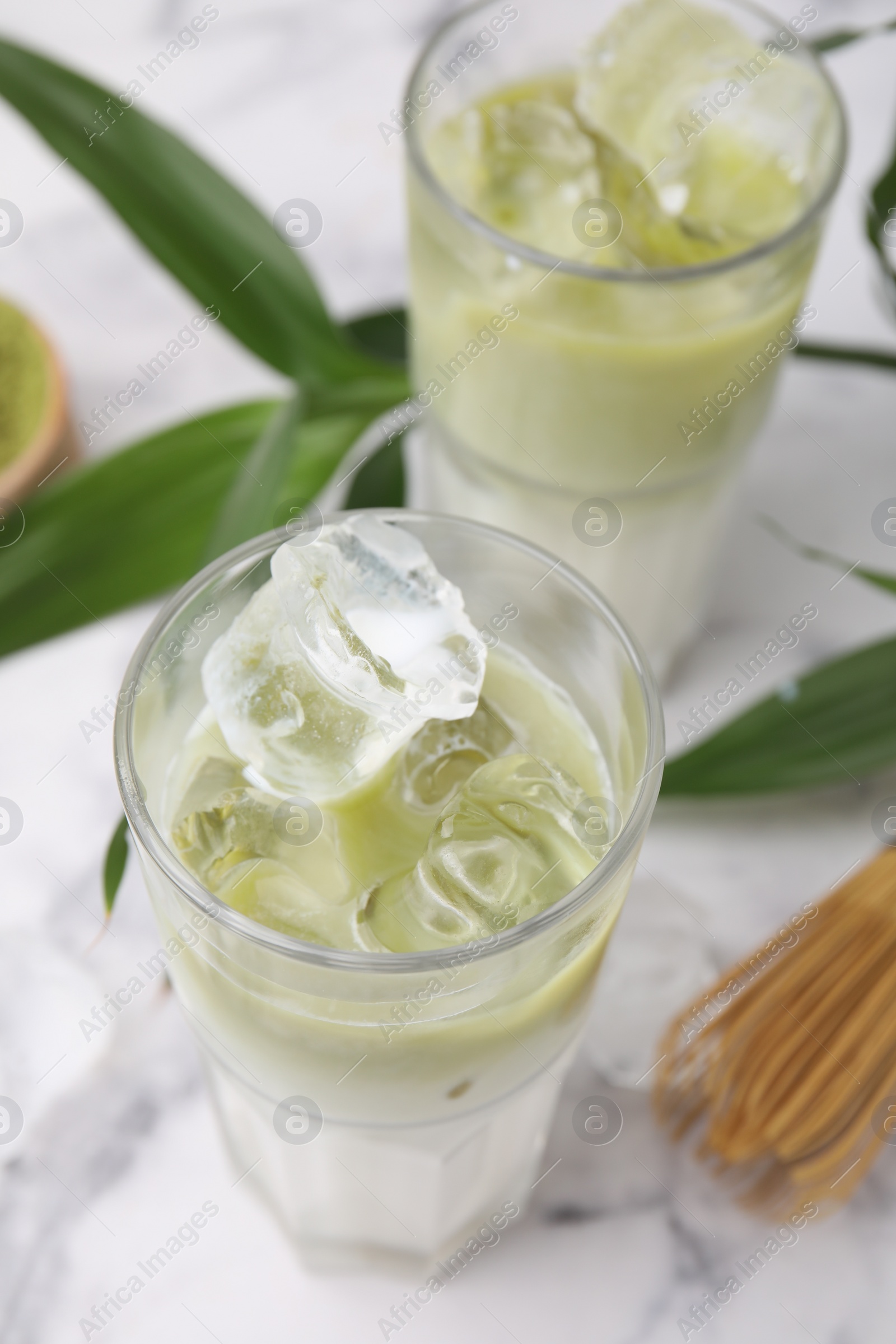 Photo of Glasses of tasty iced matcha latte, bamboo whisk and leaves on white marble table, closeup