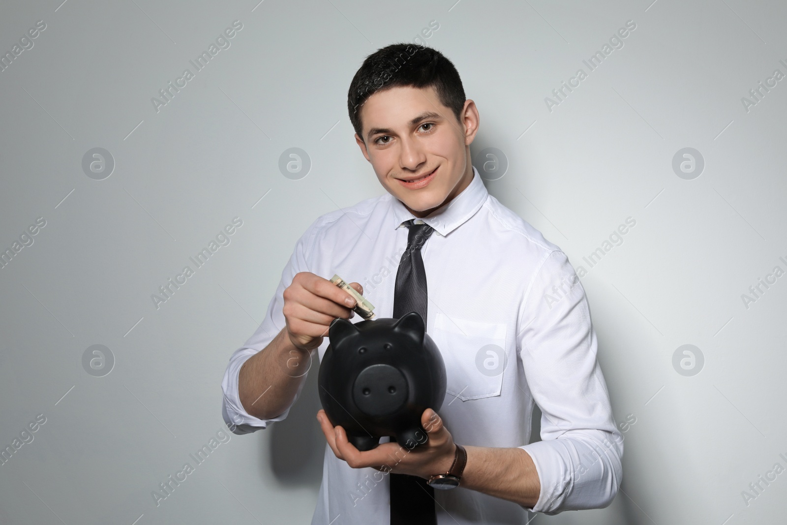 Photo of Young businessman putting money into piggy bank on light background