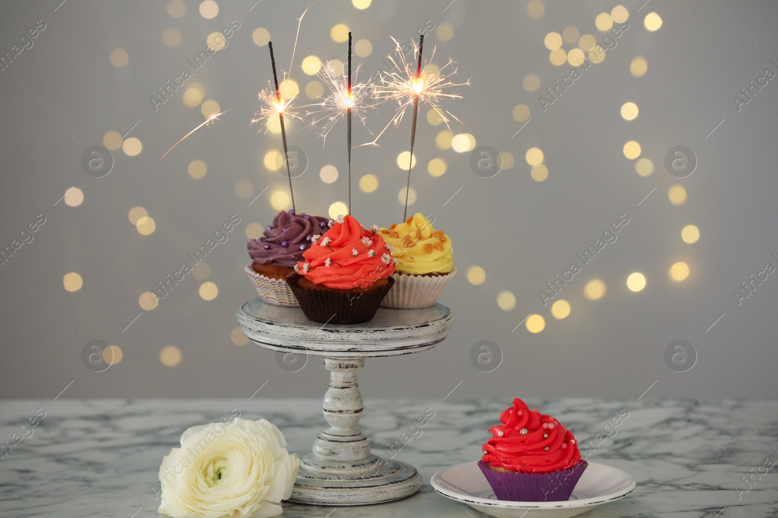 Photo of Different colorful cupcakes with sparklers on white marble table against blurred lights