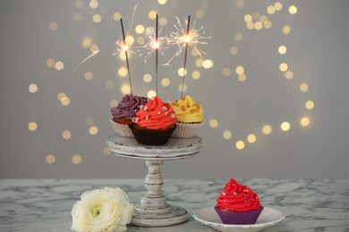 Photo of Different colorful cupcakes with sparklers on white marble table against blurred lights