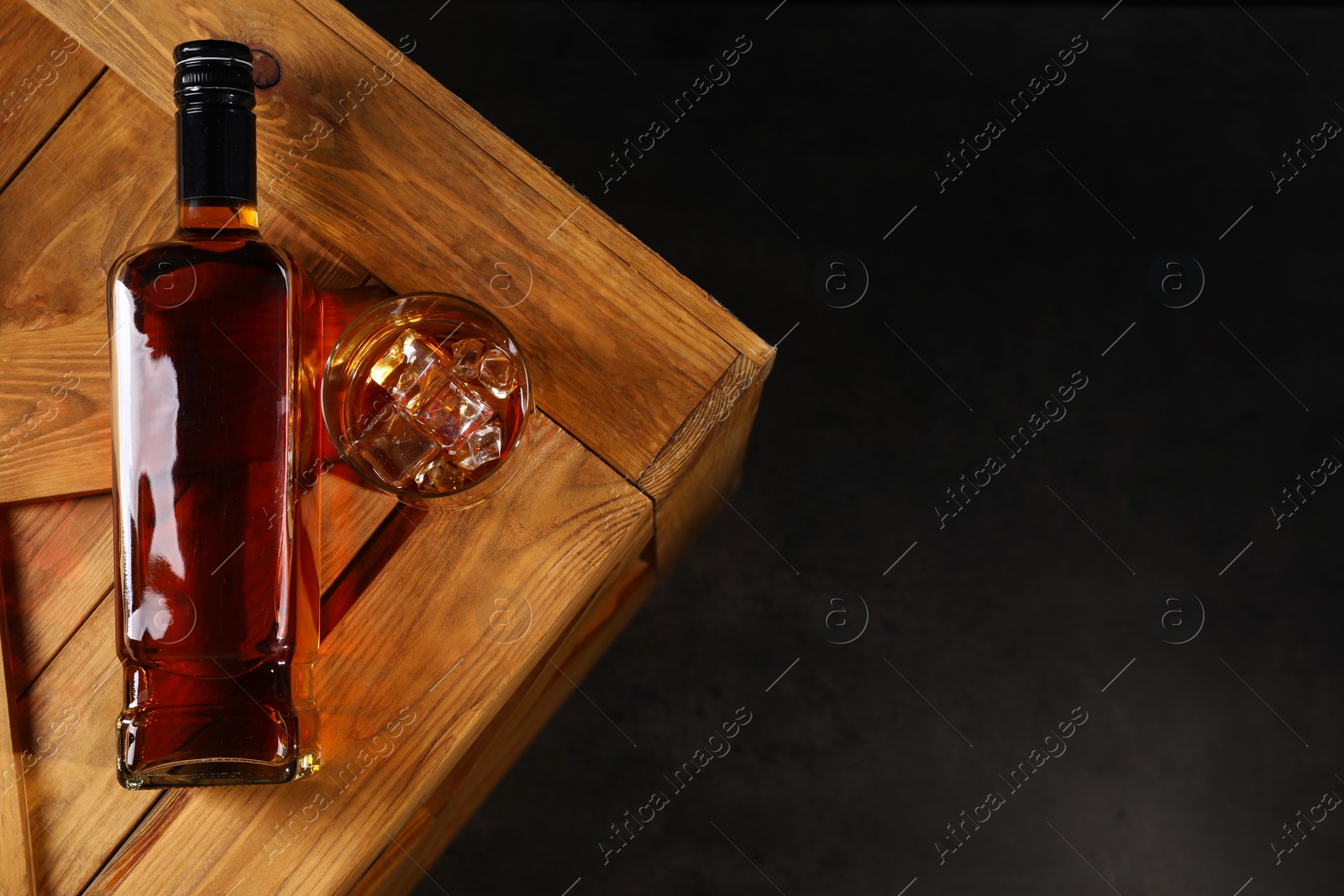 Photo of Whiskey with ice cubes in glass and bottle on wooden crate against black background, top view. Space for text