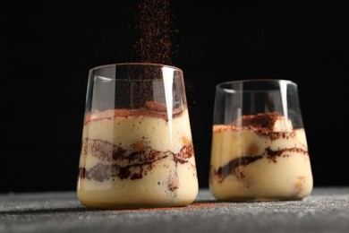 Photo of Adding cocoa powder into glass with delicious tiramisu at grey textured table, closeup