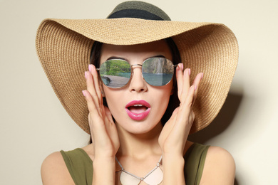 Emotional young woman wearing stylish sunglasses with reflection of lake and hat on beige background 