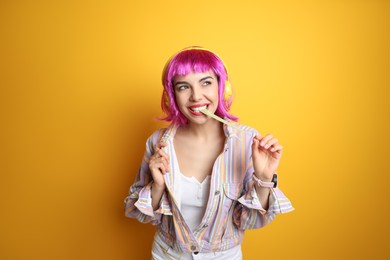Photo of Fashionable young woman in colorful wig with headphones chewing bubblegum on yellow background