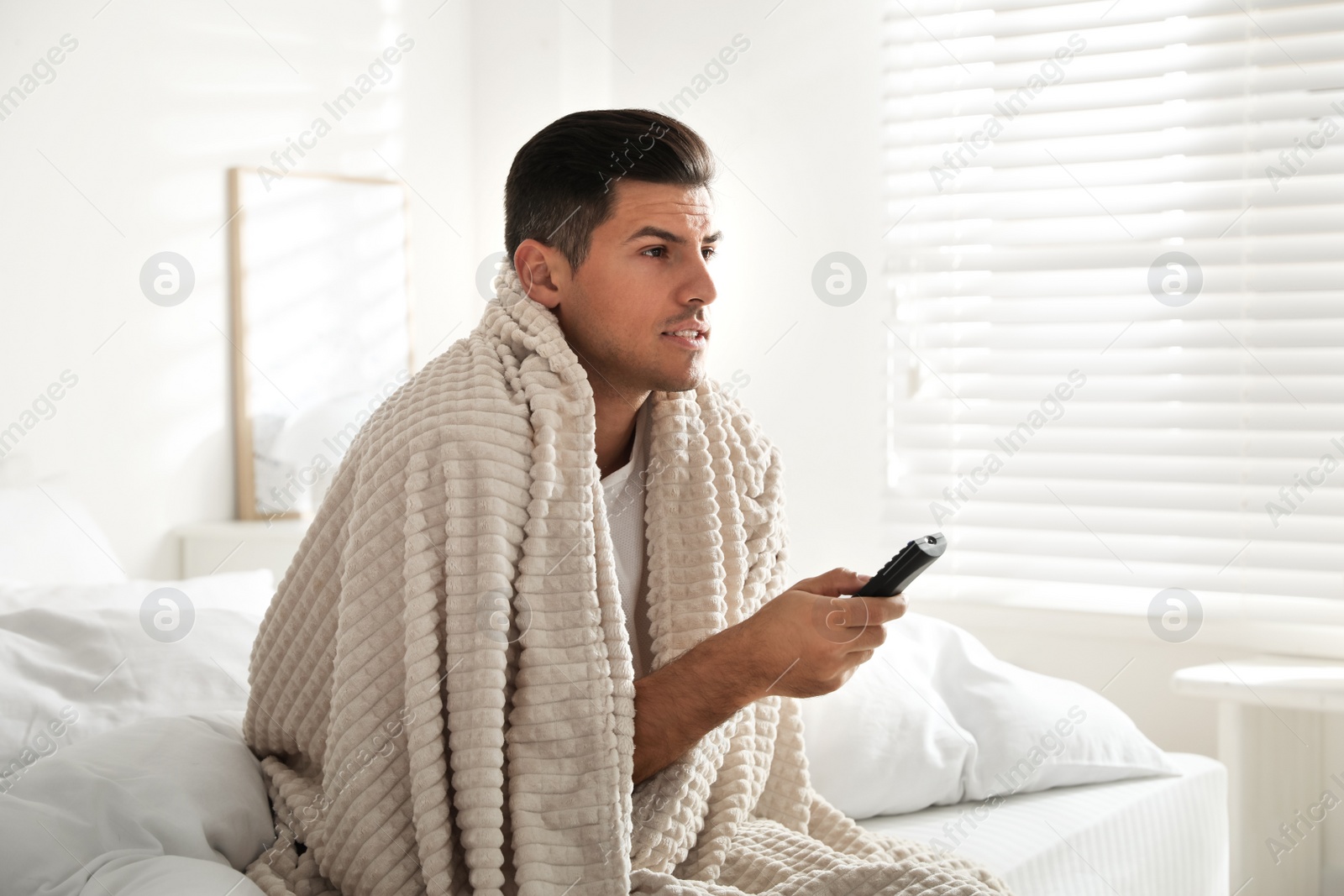 Photo of Man covered with warm beige plaid watching television in bedroom