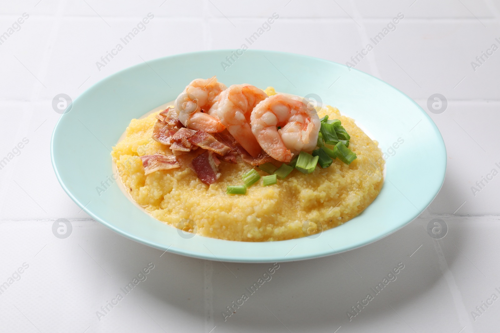 Photo of Plate with fresh tasty shrimps, bacon, grits and green onion on white tiled table