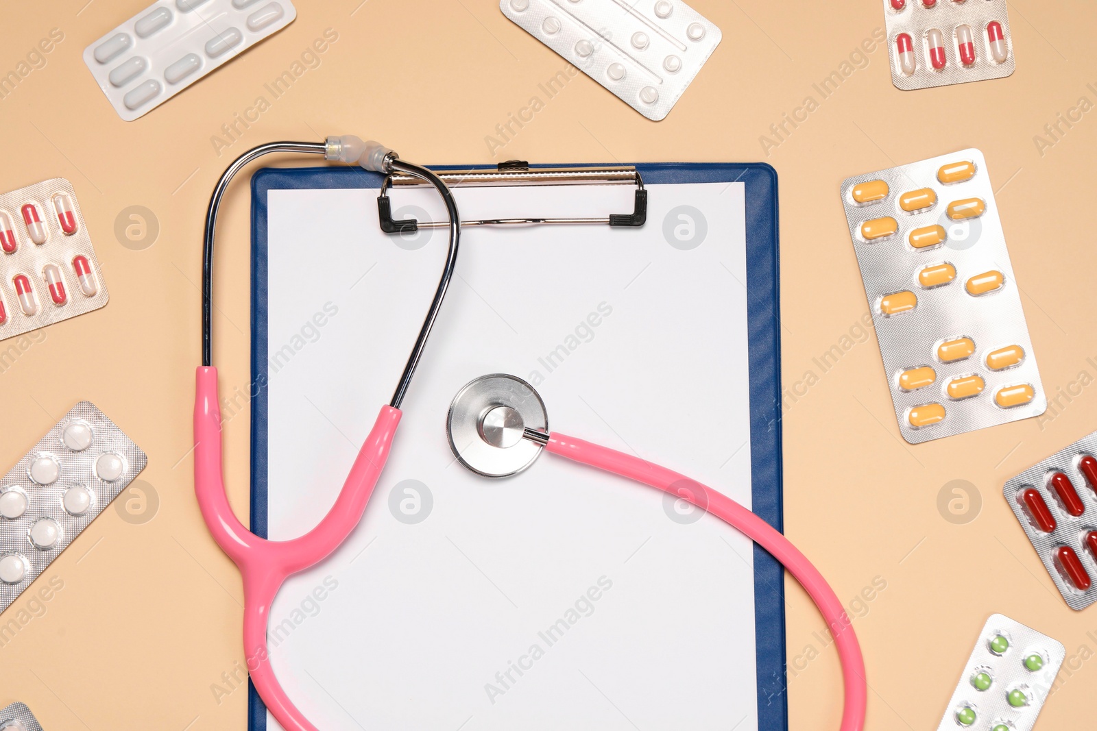 Photo of Stethoscope and clipboard surrounded by pills on beige background, flat lay. Medical tool