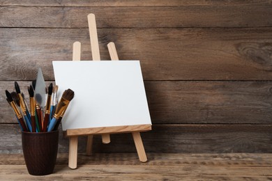 Photo of Easel with blank canvas and brushes on wooden table. Space for text