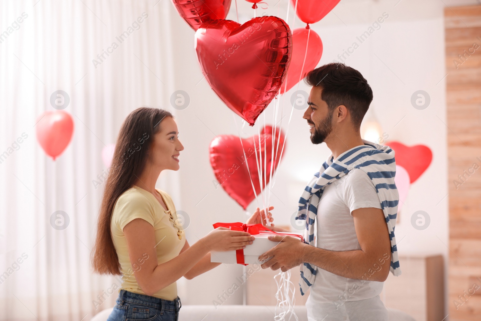 Photo of Lovely young couple with gift box and heart shaped  balloons in living room. Valentine's day celebration