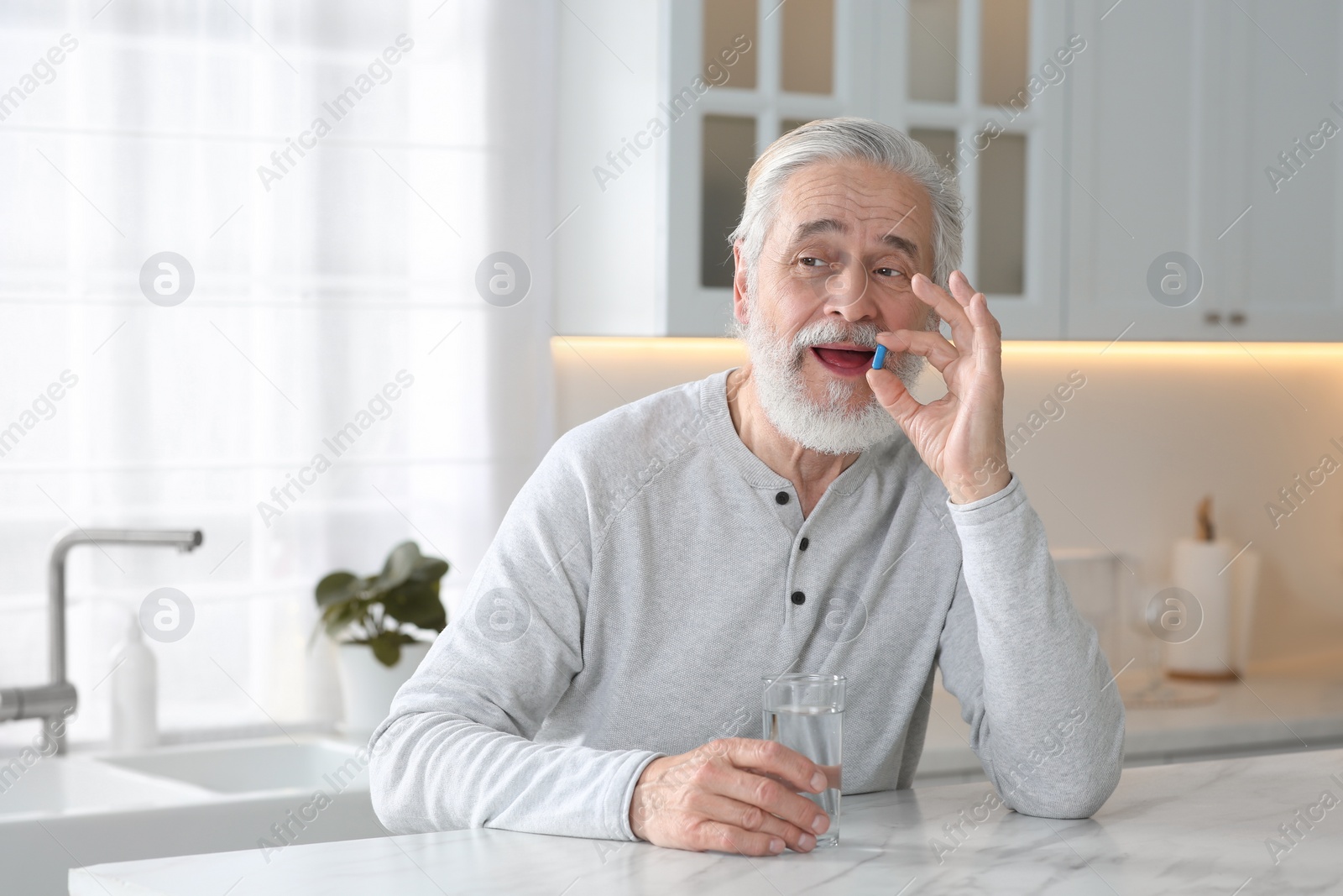 Photo of Senior man with glass of water taking pill at table in kitchen