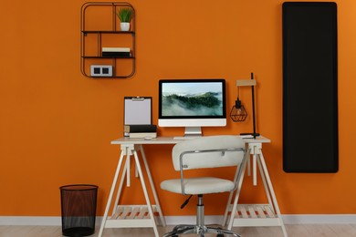 Workplace with modern computer on wooden desk and comfortable chair near orange wall. Home office