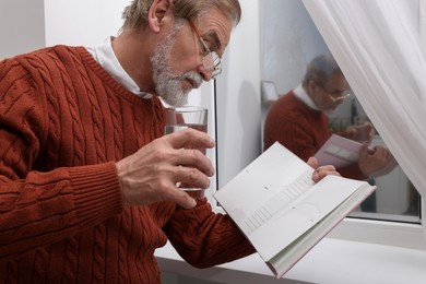 Photo of Upset senior man with glass of water reading book near window at home. Loneliness concept