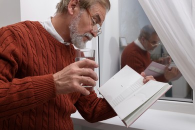 Upset senior man with glass of water reading book near window at home. Loneliness concept