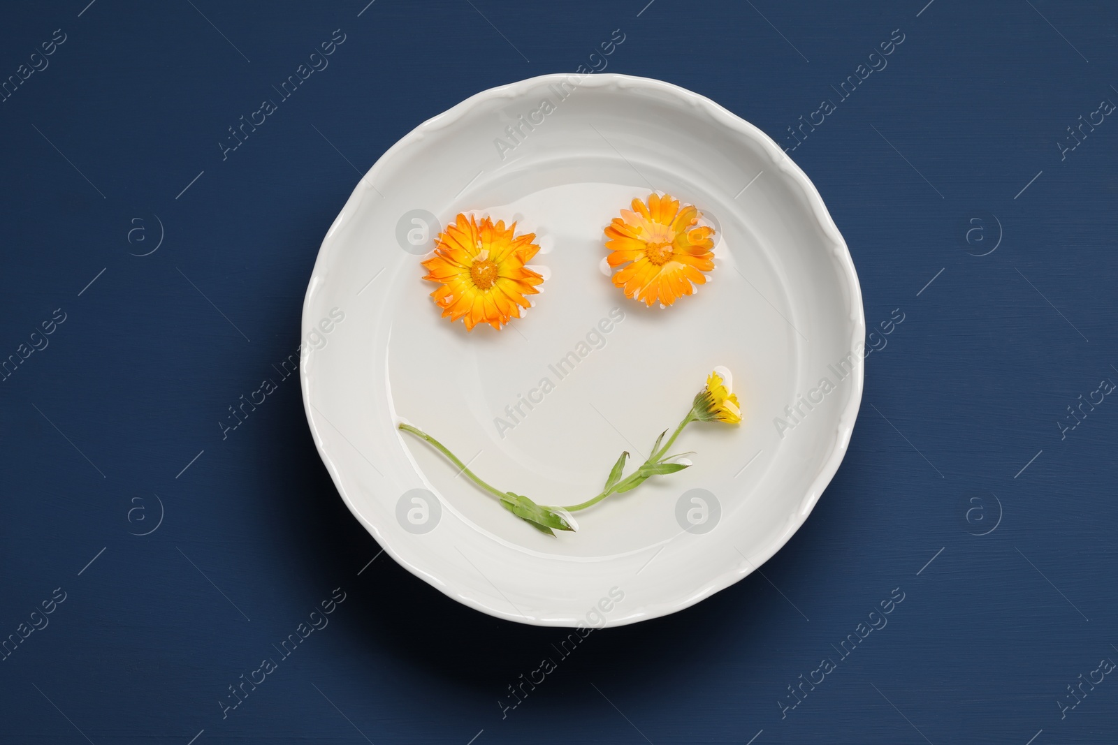 Photo of Face with smile made of beautiful fresh calendula flowers in bowl of water on blue wooden table, top view