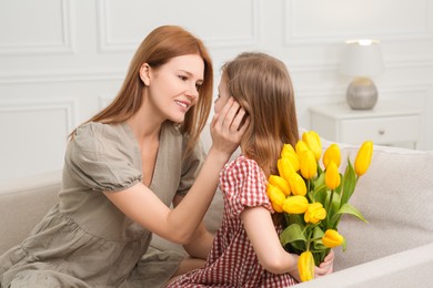 Photo of Cute daughter hiding bouquet of yellow tulips for her mother on sofa at home