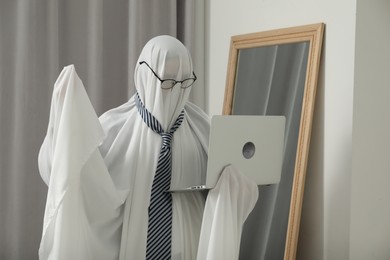 Photo of Creepy ghost. Man in white sheet using laptop at home